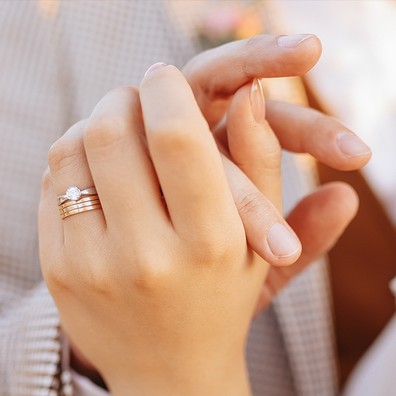 2 hands, one with an engagement ring and stackable rings on one finger, interconnected